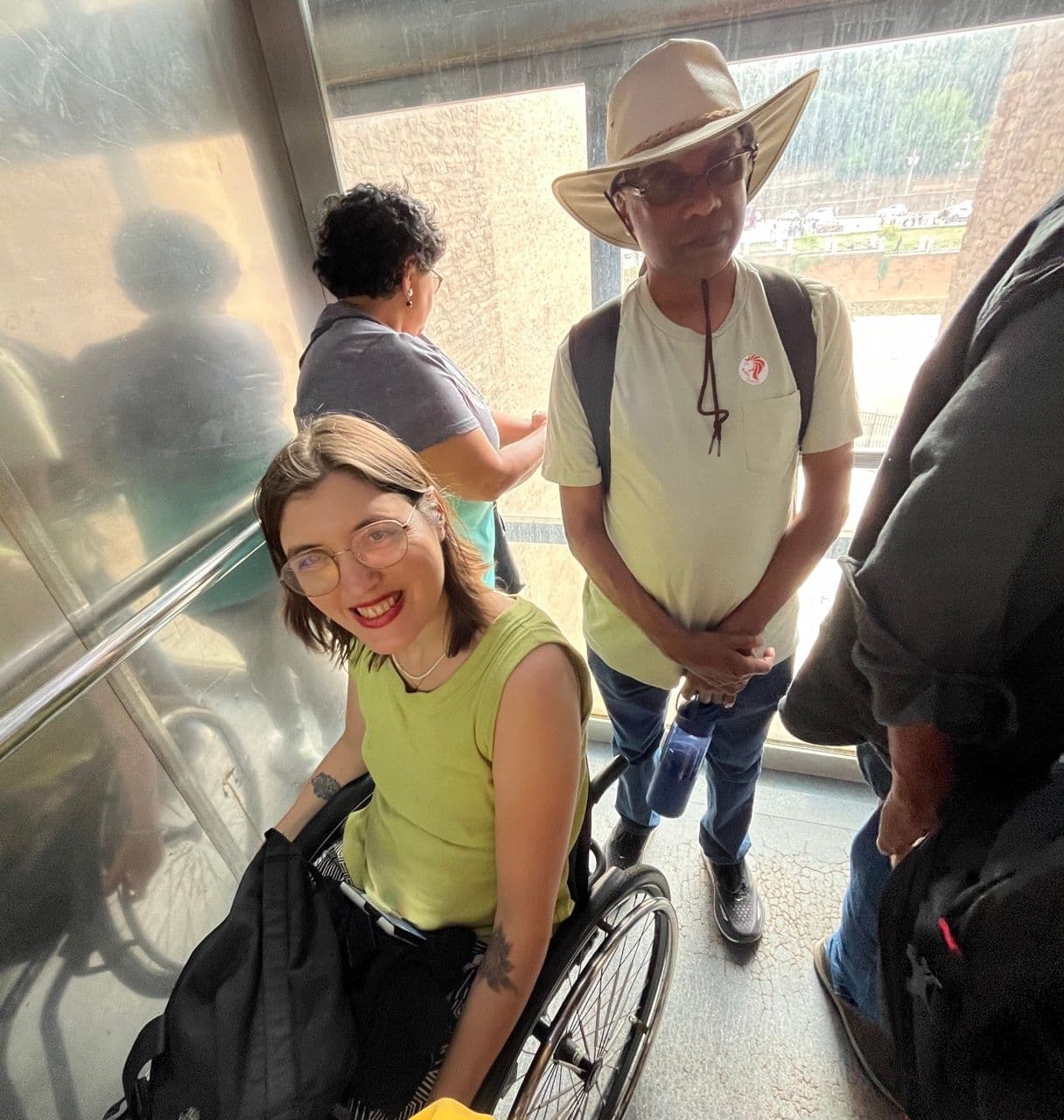 Mechtihild, a young woman on a manual wheelchair smiling inside the elevator of the Coliseum with two other people