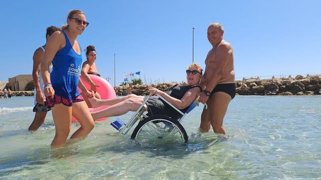 A lady on a beach wheelchair is carried in the sea by smiling volunteers